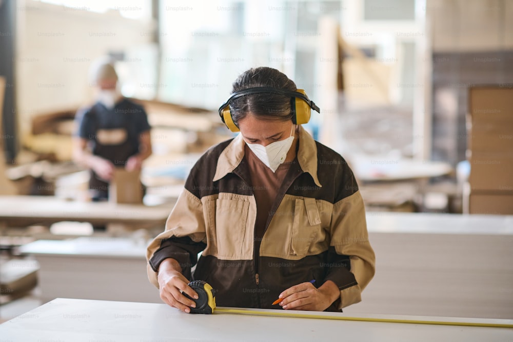 Junge Arbeiterin mit Schutzmaske und Kopfhörer misst mit Maßband auf Holzbrettern