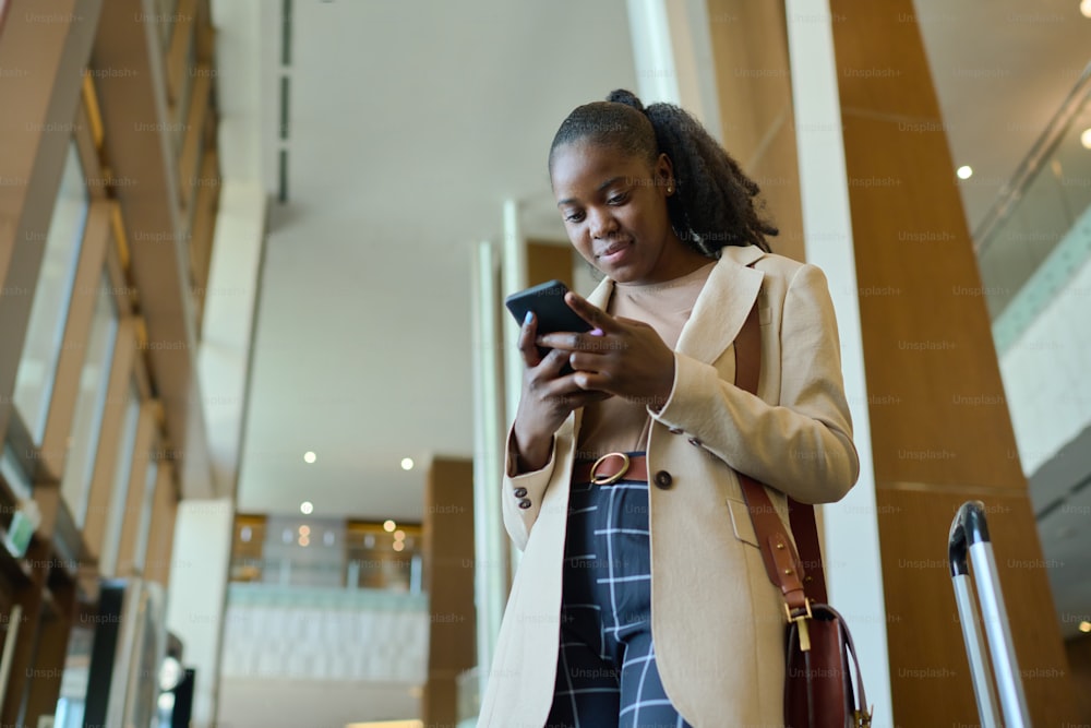 Young elegant African American businesswoman with mobile phone talking through video chat while standing in hotel lounge