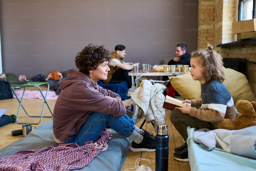 Young woman in casualwear listening to her little son with open book reading it and discussing with his mother in room for refugees