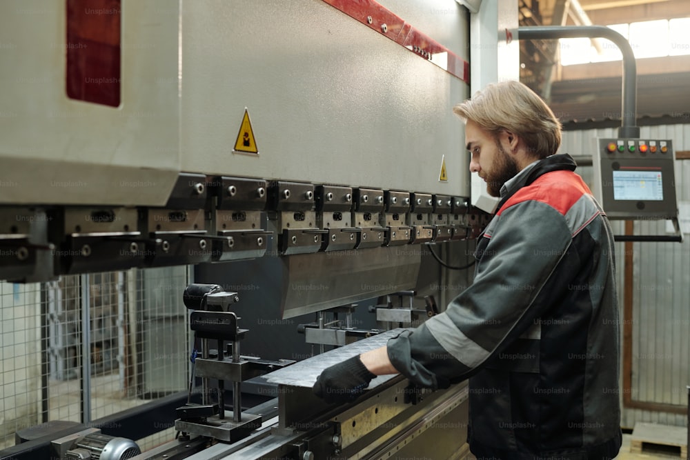 Jovem engenheiro do sexo masculino em pé na frente da enorme máquina industrial e verificando a qualidade das novas chapas metálicas enquanto trabalha na fábrica