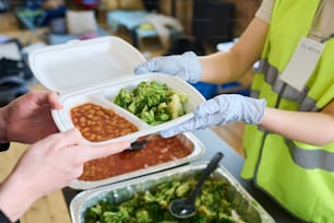 Gloved hands of female volunteer passing container with cooked food to boy from refugee camp or shelter for temporarily homeless people