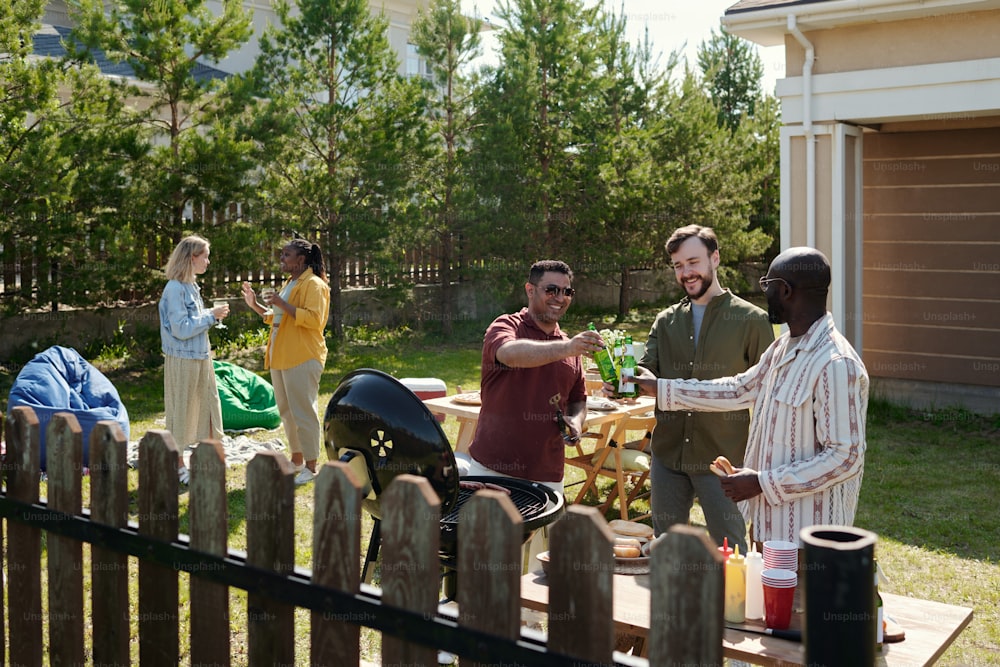 Three happy young intercultural men clinking with bottles of beer during outdoor party on weekend while one of them making barbecue on grill