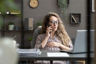 Young pregnant businesswoman in eyeglasses talking on mobile phone while sitting at her workplace with laptop at office