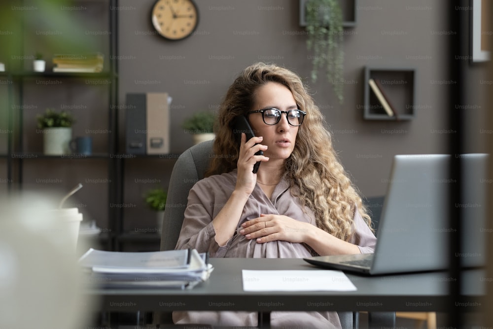 Jovem empresária grávida de óculos falando ao telefone celular enquanto está sentada em seu local de trabalho com laptop no escritório