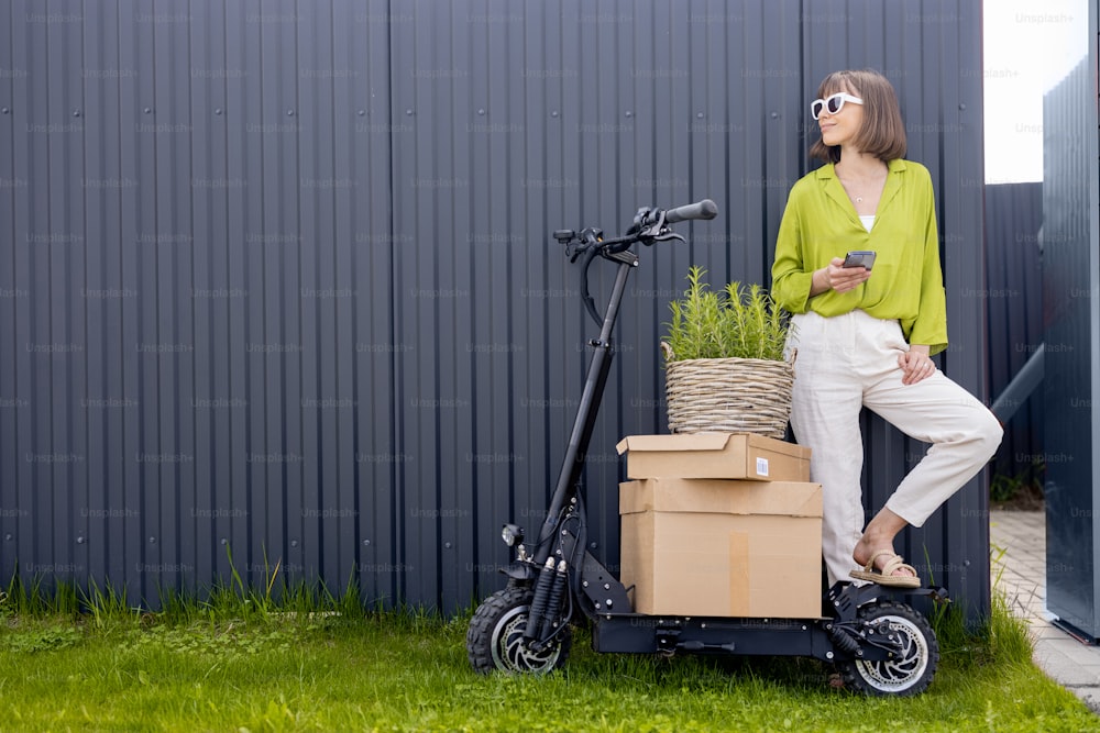 Young woman standing with phone near her electric scooter with parcels and flowerpot on gray wall background. Concept of delivery and sustainable lifestyle