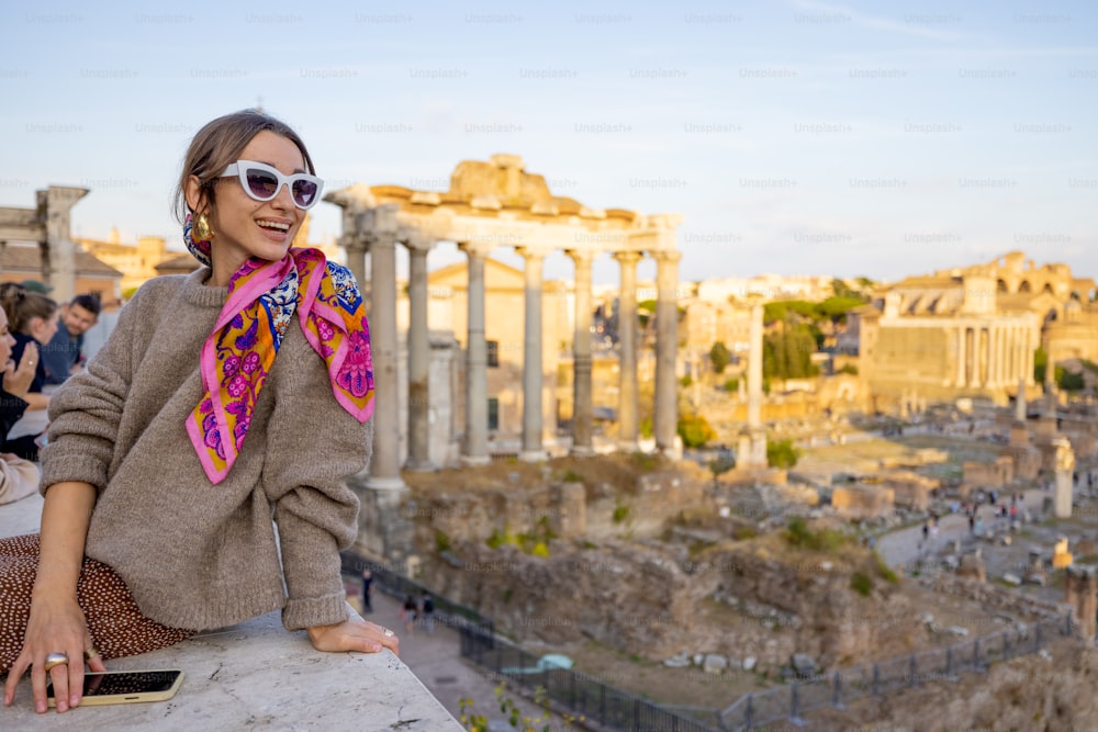 Portrait of a cheerful woman in front of the Roman Forum, ruins at the center of Rome on a sunset. Concept of traveling famous landmarks in Italy. Caucasian woman wearing colorful shawl and sunglasses