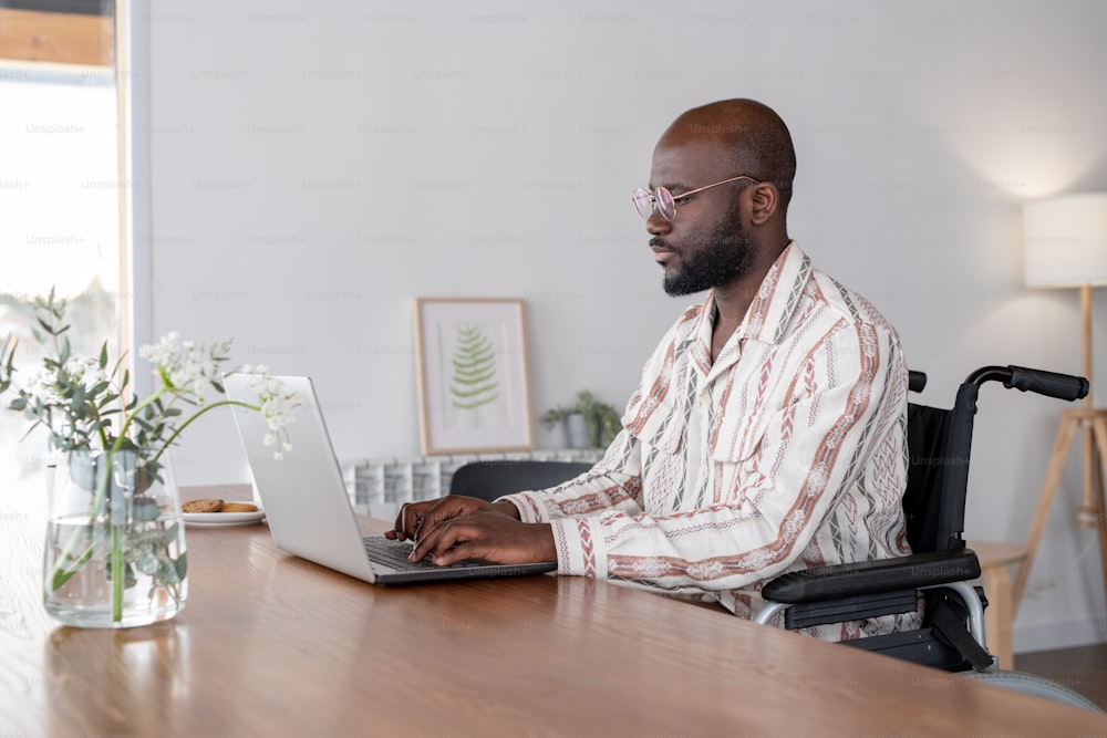 Joven hombre de negocios serio o autónomo en silla de ruedas sentado junto a la mesa en la sala de estar y mirando la pantalla de la computadora portátil mientras escribe