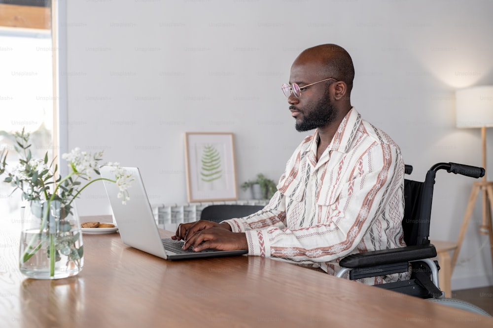 Jeune homme d’affaires sérieux ou pigiste en fauteuil roulant assis près d’une table dans le salon et regardant l’écran d’un ordinateur portable tout en tapant