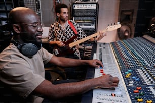 Jeune musicien noir moderne jouant de la guitare électrique tout en utilisant une console de mixage dans un studio d’enregistrement