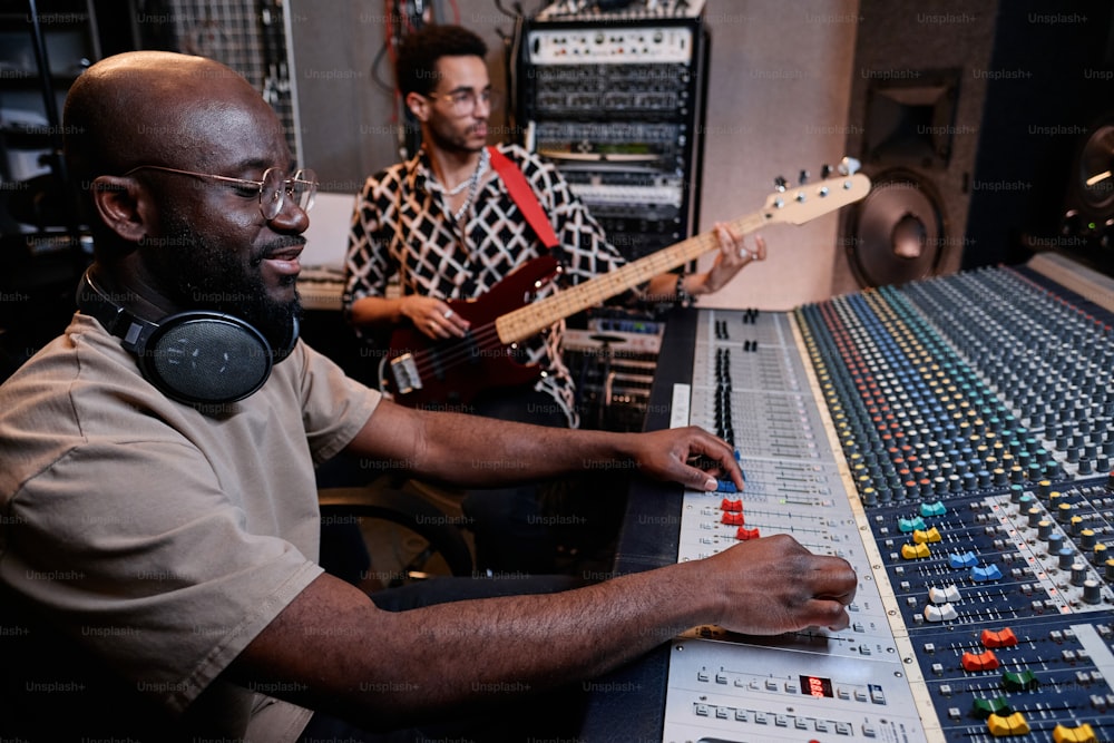 Modern young Black musician playing electric guitar while sound engineer using mixing console in recording studio
