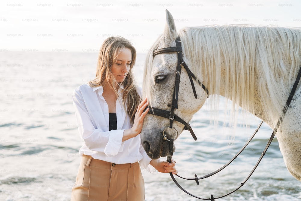 Junge langhaarige Frau im weißen Hemd mit weißem Pferd auf Seelandschaftshintergrund