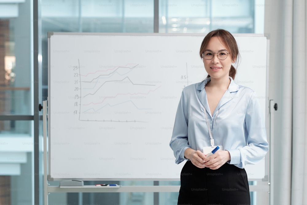 Jovem professor bem-sucedido de economia em roupas formais de pé ao lado do quadro branco com gráficos durante a apresentação dos dados