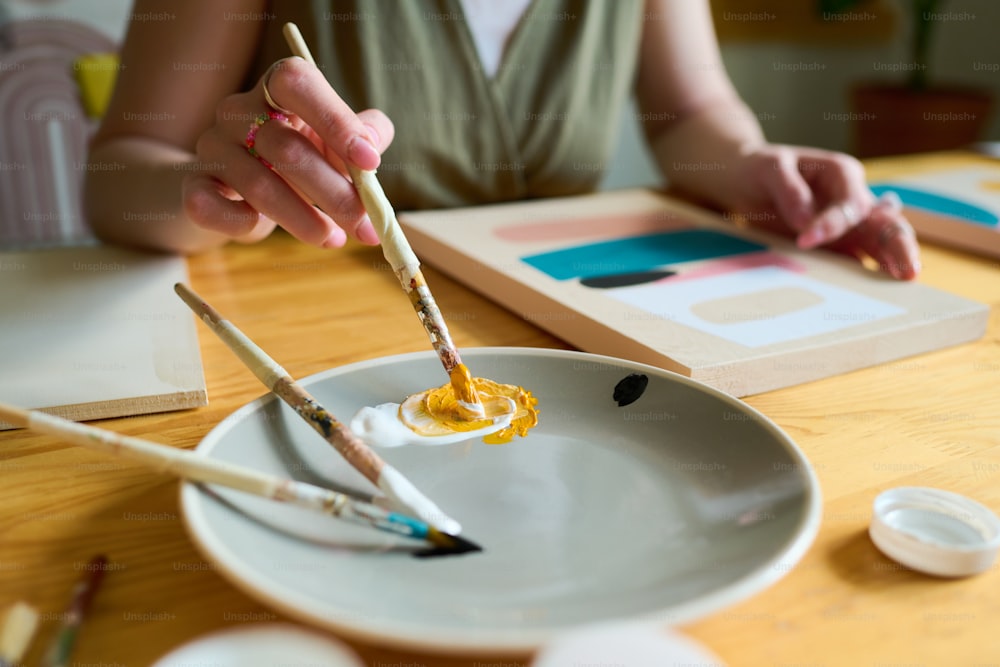 Hand einer jungen kreativen Frau mit Pinsel, die weiße und gelbe Farben auf dem Teller mischt, während sie am Tisch sitzt und auf Papier malt