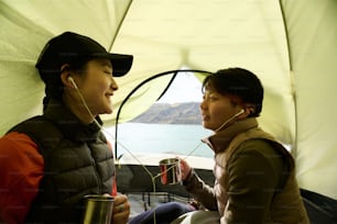 Dos alegres jóvenes asiáticas sentadas juntas en una tienda de campaña escuchando música con auriculares y bebiendo té o café