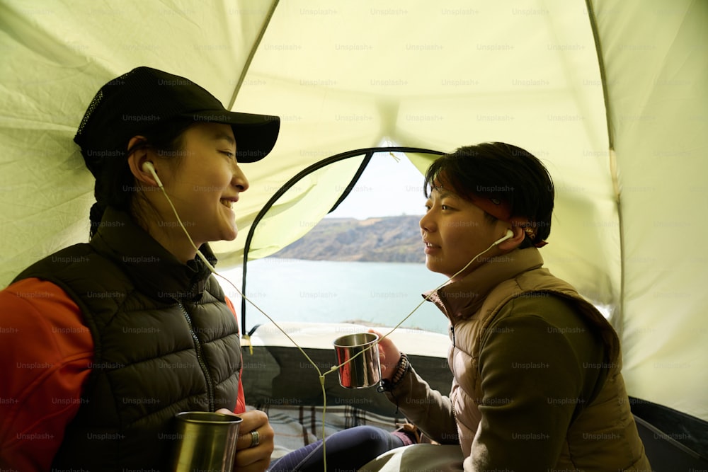 Two joyful young Asian women sitting together in tent listening to music in earphones and drinking tea or coffee