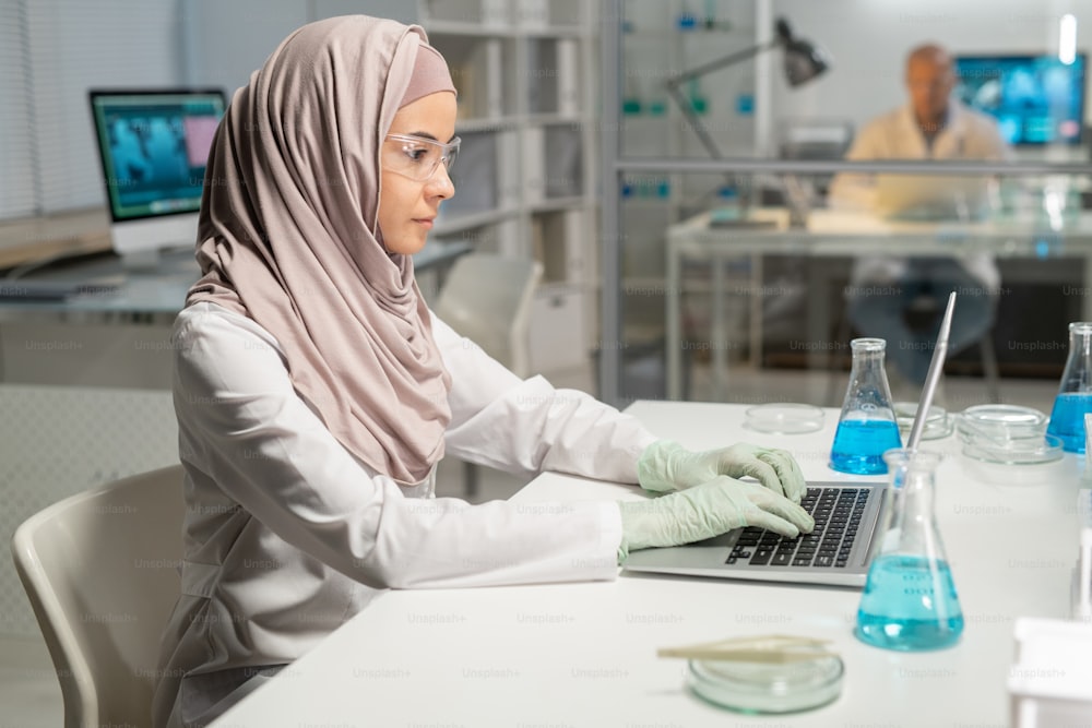 Mulher muçulmana nova no hijab, jaleco, óculos de proteção e luvas sentada na mesa na frente do laptop no laboratório e digitação