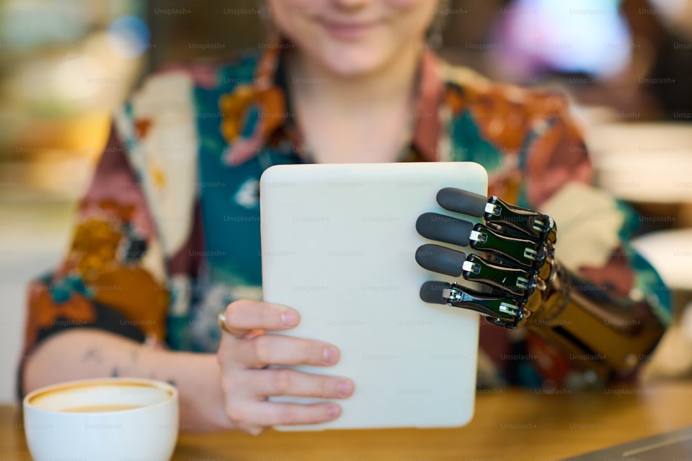 Hände einer jungen Frau mit myoelektrischem Arm, die das Tablet vor sich hält, während sie am Tisch im Café sitzt und Kaffee trinkt