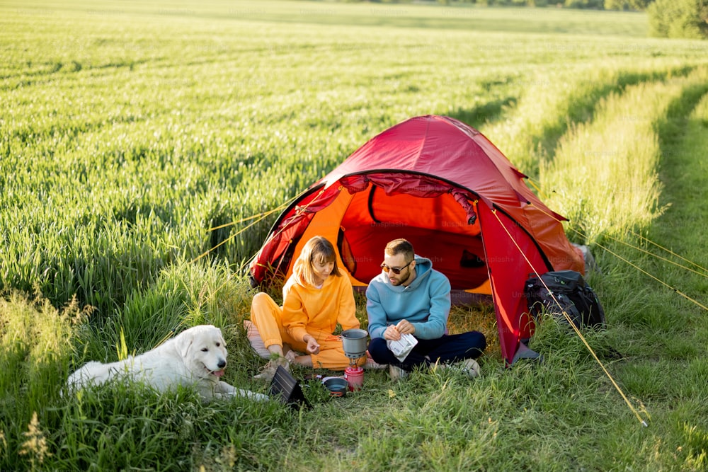 Young couple have a picnic near the tent, while traveling with dog on nature. Man and woman spending summer time with pet camping on greenfield on summer evening