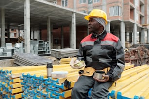 Restful mature builder with snack and cup of tea sitting on stack of yellow building materials and having lunch at construction area