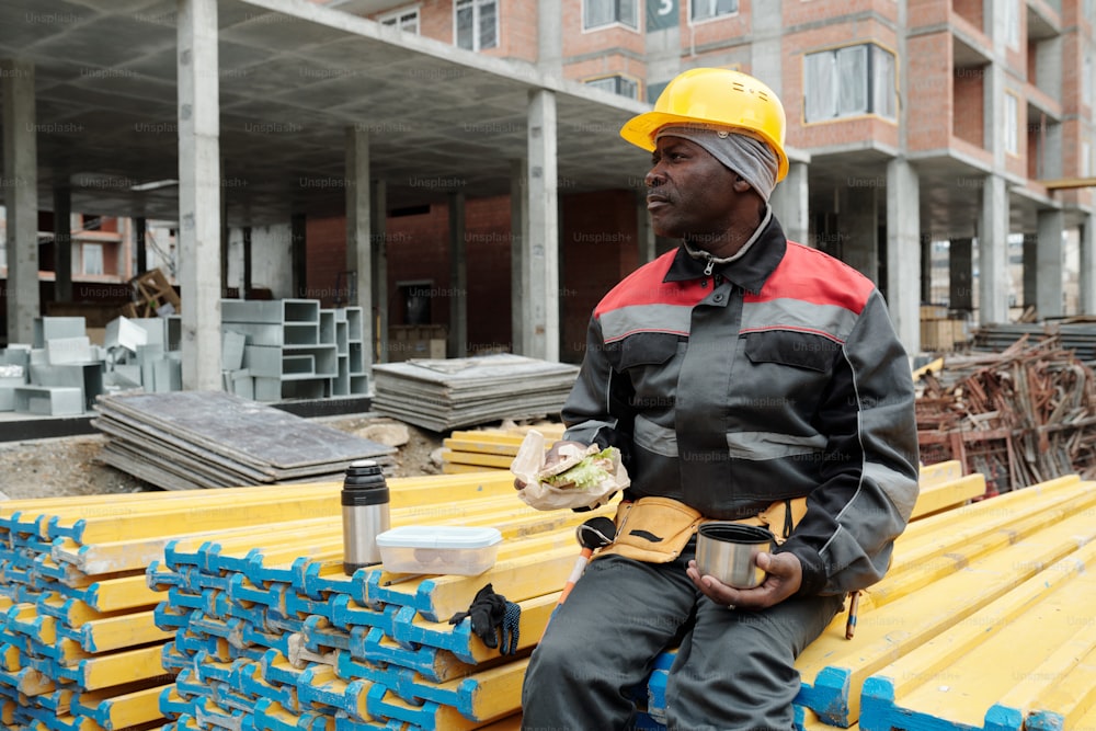 Construtor maduro repousante com lanche e xícara de chá sentado na pilha de materiais de construção amarelos e almoçando na área de construção