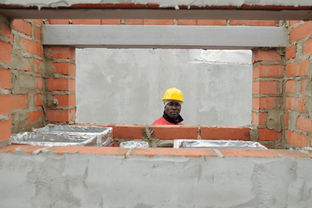보호 헬멧과 작업복을 입은 성숙한 아프리카계 미국인 건축업자는 창틀과 건축 자재가 있는 벽돌 벽 옆에 서 있습니다.