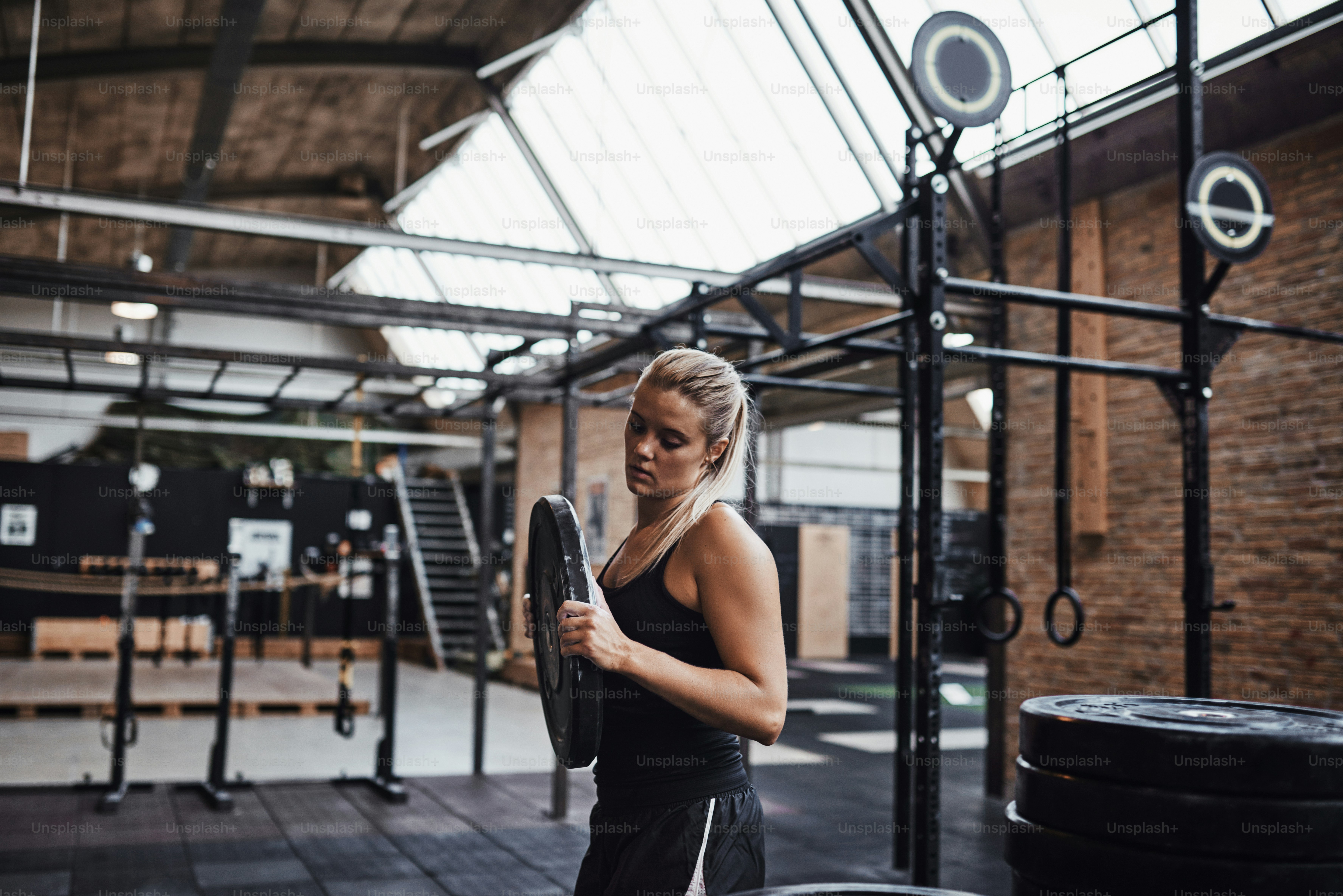 Young blonde woman in sportswear selecting weights for a workout session while standing alone in a gym