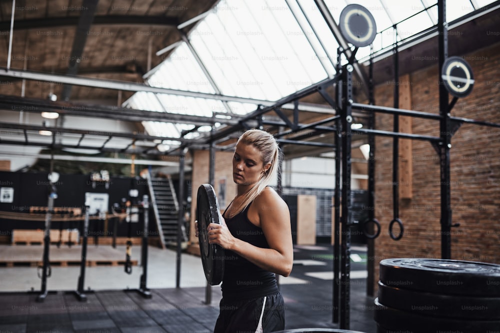 Mulher loira jovem em roupas esportivas selecionando pesos para uma sessão de treino enquanto está sozinha em uma academia