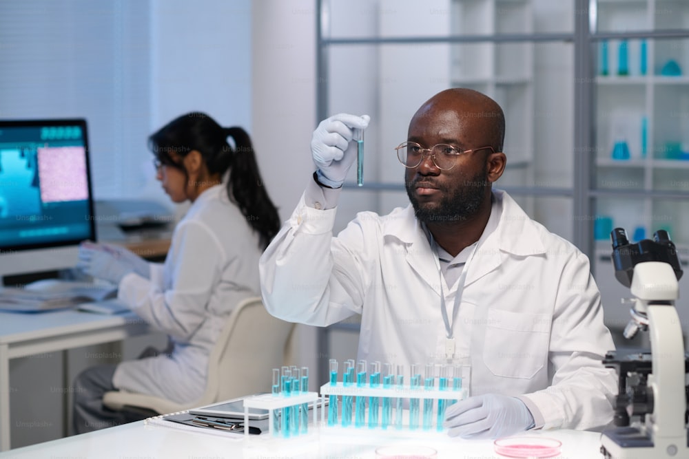 Afroamerikanischer männlicher Virologe in Handschuhen und Laborkittel schaut auf blaue Flüssigkeit in Flasche gegen Kollegin, die vor dem Computer arbeitet