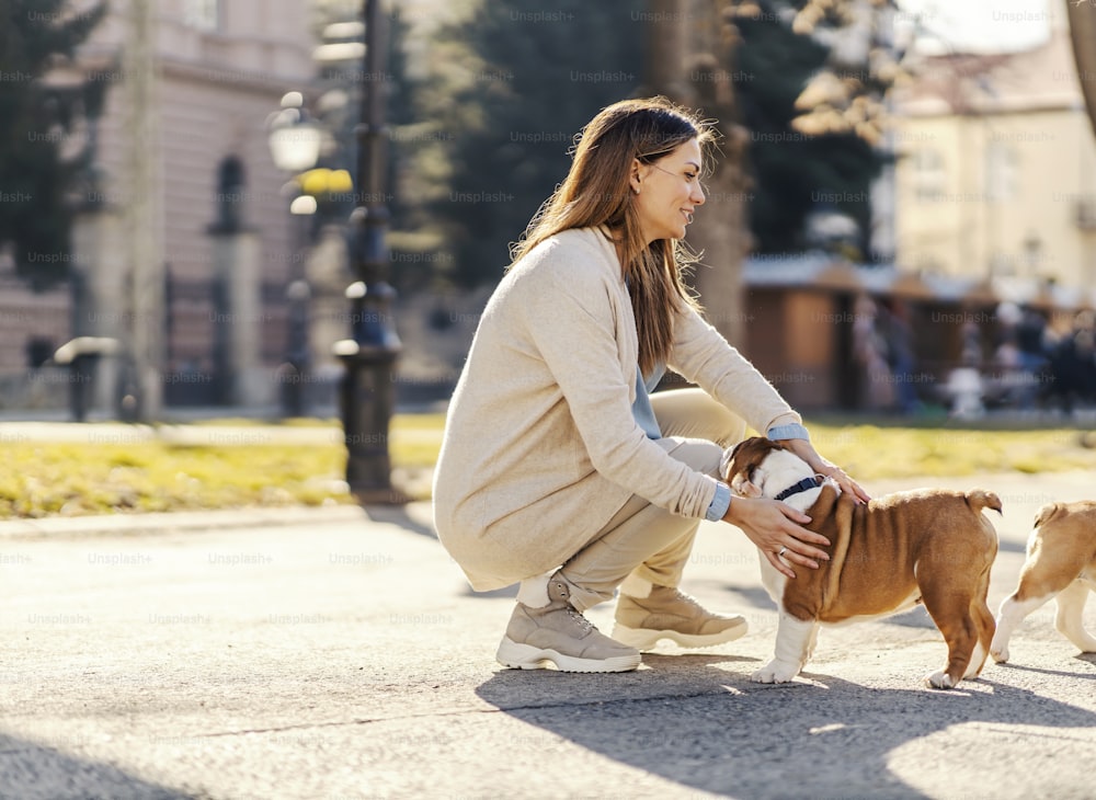 A smiling woman crouching in a park and petting bulldog on a leash.