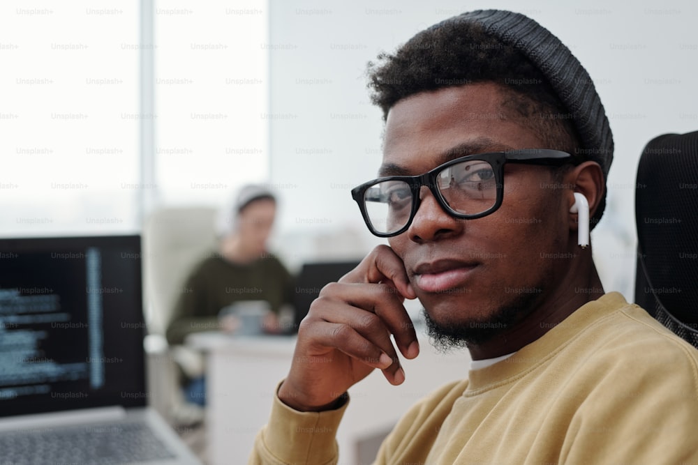 Visage d’un jeune programmeur afro-américain sérieux portant des lunettes, des vêtements décontractés et des écouteurs regardant la caméra près de son lieu de travail