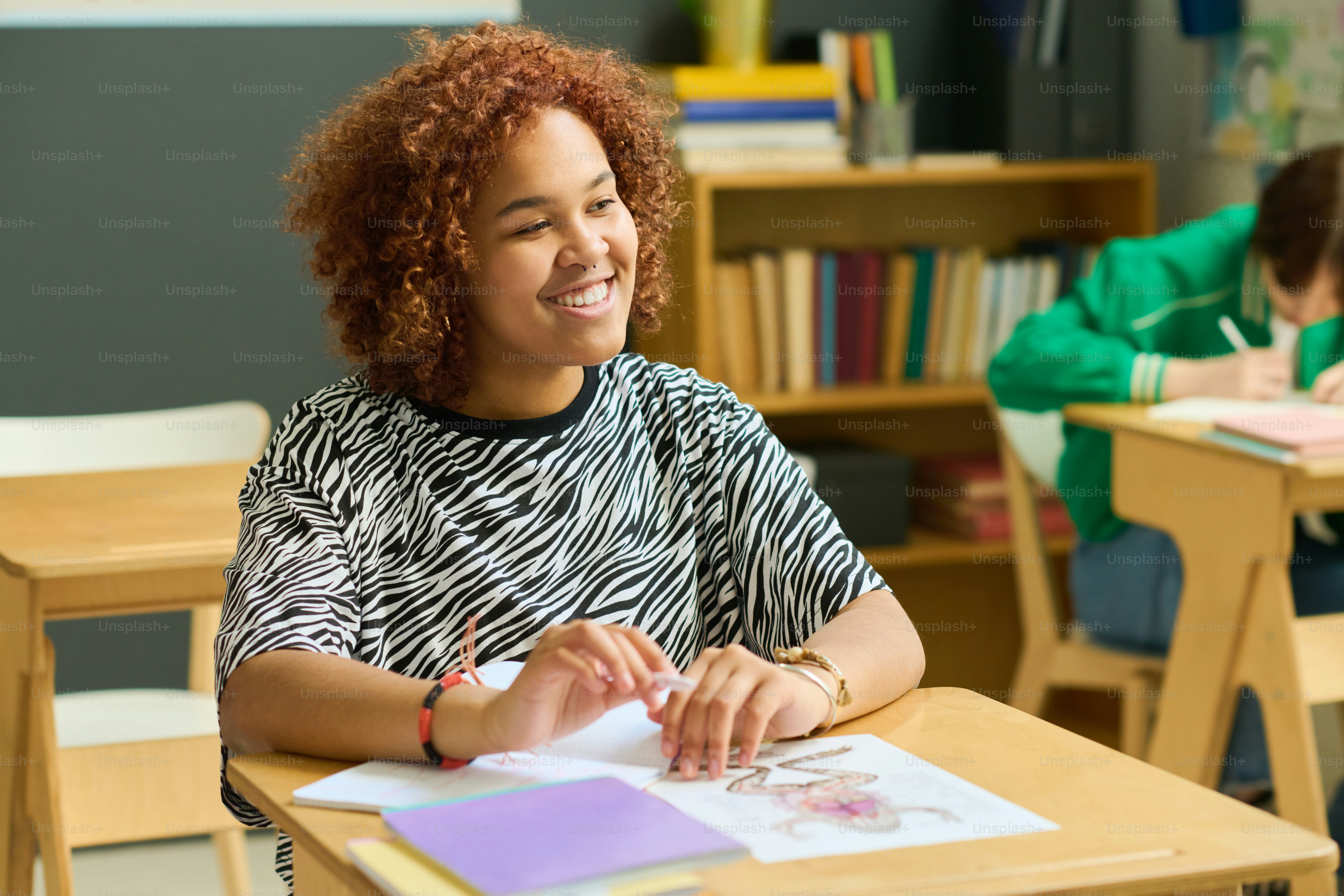 Happy female student of high school sitting by desk and looking at teacher with smile while listening to explanation of new subject