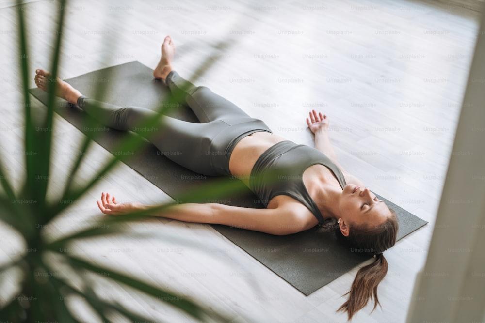 La giovane donna in forma pratica lo yoga facendo savasana nello studio di yoga leggero con la pianta della serra