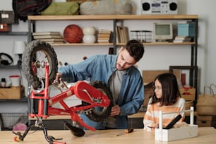 Joven hombre contemporáneo revisando las ruedas de la bicicleta de su hija en el garaje mientras ambos están de pie junto a la mesa de madera con herramientas de mano