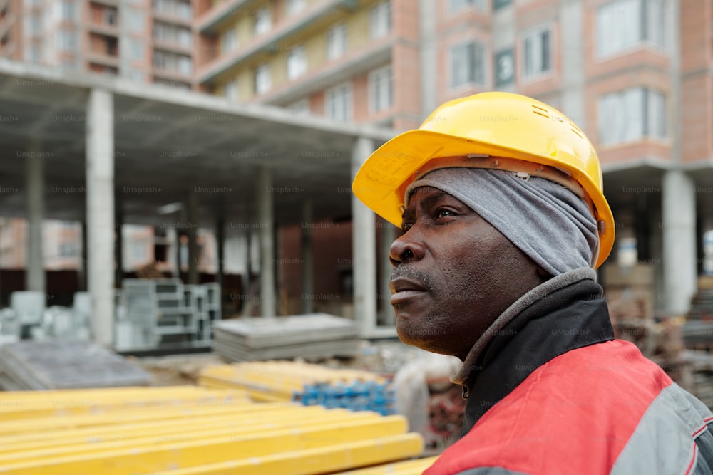 Afroamerikanischer männlicher Bauarbeiter in Arbeitskleidung und Schutzhelm, der zur Seite schaut, während er auf der Baustelle gegen ein unfertiges Gebäude steht