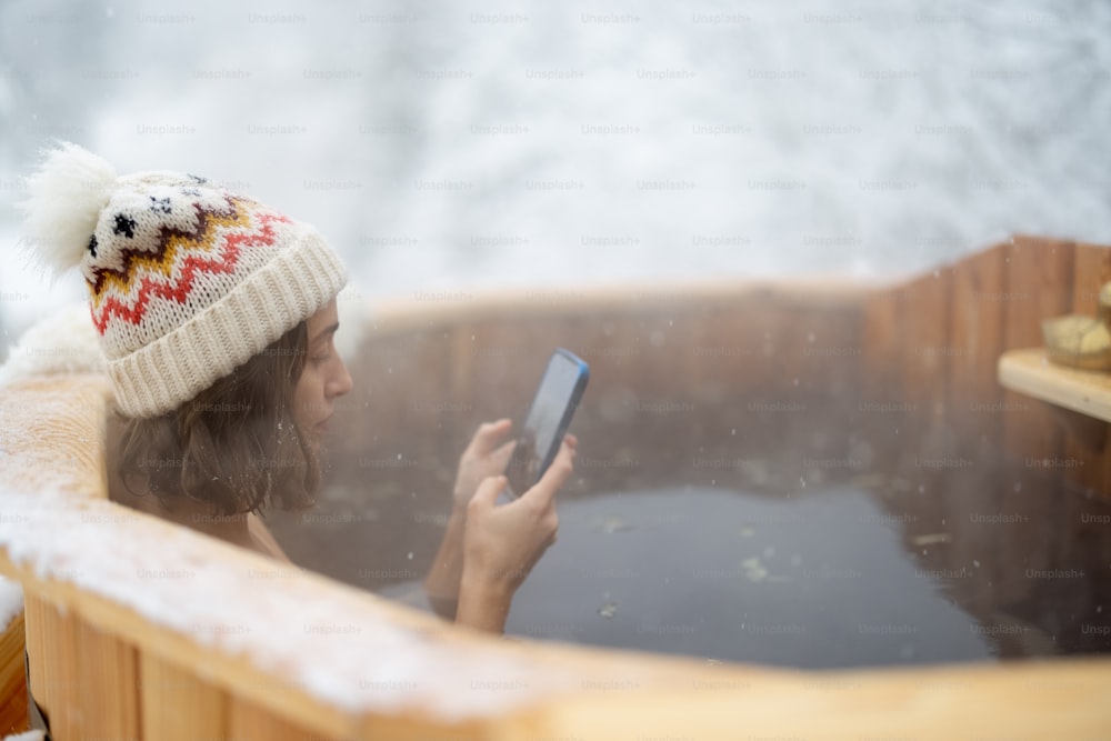 Woman relaxing in hot bath outdoors, enjoying thermal spa at snowy mountains. Winter recreation and water treatments concept. Caucasian woman in hat with smart phone