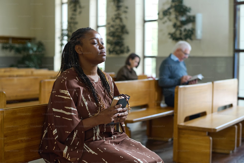 캐주얼 드레스를 입은 젊은 기독교 흑인 여성이 교회 벤치에 앉아 주님께 기도하는 동안 눈을 감고 있다.