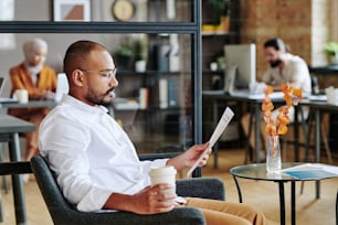 Side view of serious young male economist looking through financial document and having coffee while sitting in armchair by small table