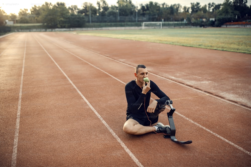 Um corredor com prótese de perna relaxando com música e comendo maçã no estádio.