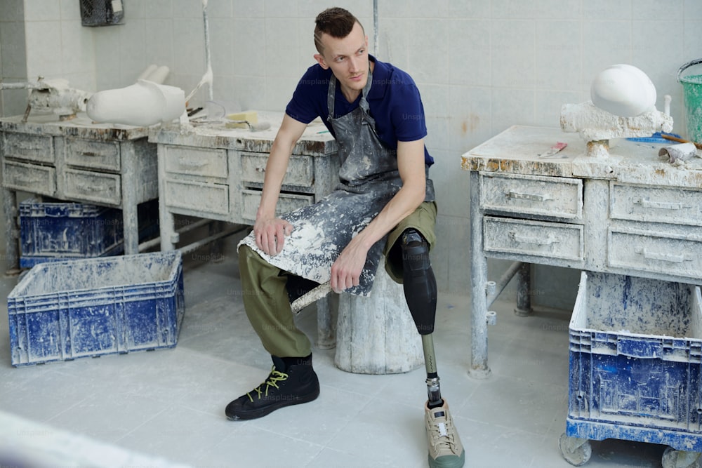 Jovem homem sério com deficiência usando avental sentado no local de trabalho e tendo descanso após o trabalho na oficina da fábrica de próteses