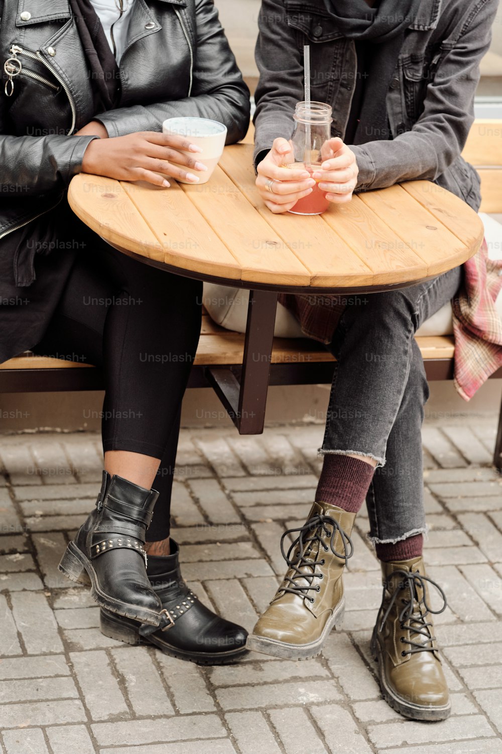 Two young intercultural females in casualwear having drinks by round wooden table outdoors