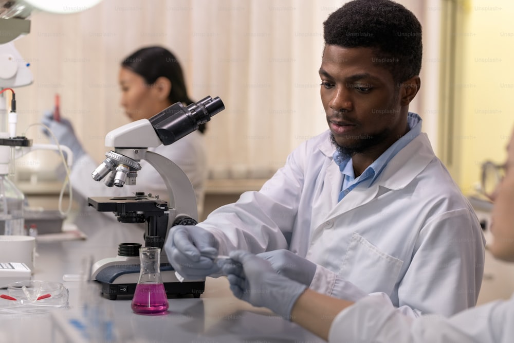 Joven científico masculino enguantado de etnia africana haciendo un experimento científico con un colega