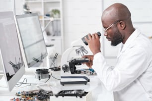 Vista laterale di giovane scienziato africano in camice bianco che studia le caratteristiche del microchip al microscopio
