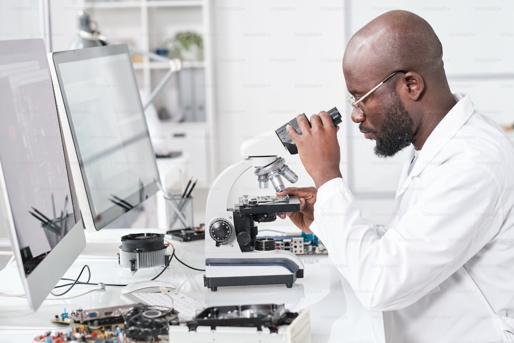 Vista lateral de un joven científico africano en bata blanca que estudia las características del microchip en el microscopio
