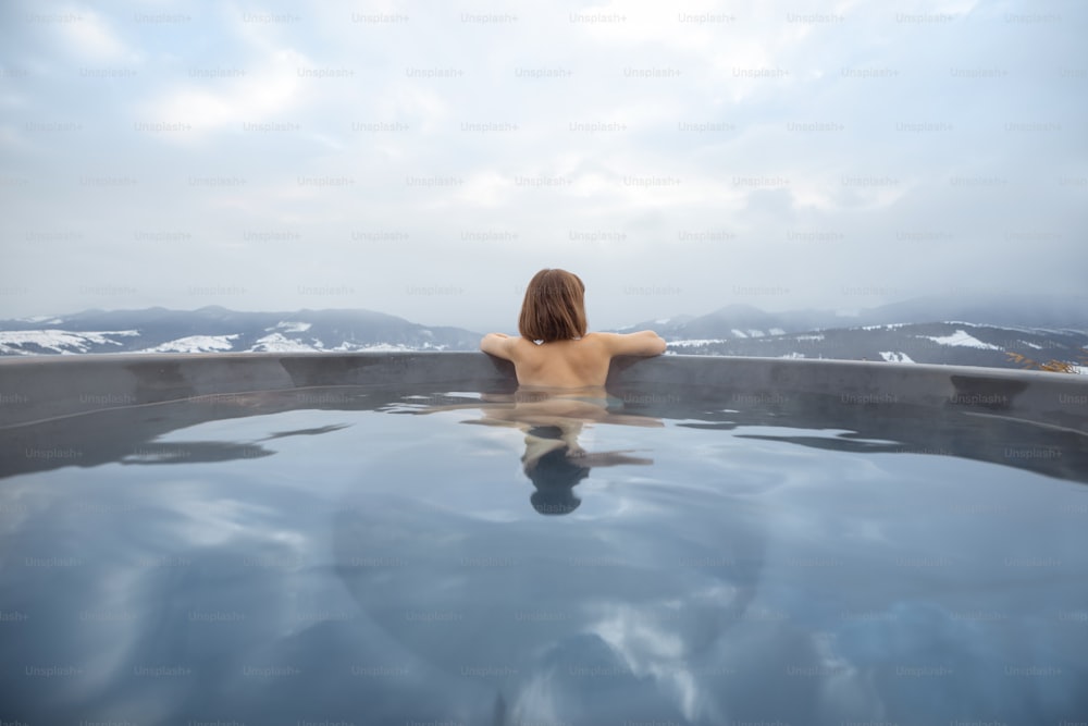 冬の山でお風呂に入浴する若い女性。自然の熱い大桶での休息と回復のコンセプト。山での逃避行とレクリエーションのアイデア