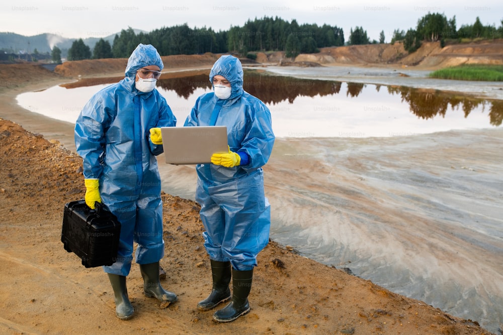 Two young female ecologists in protective workwear standing on polluted soil or clay against lake with dirty water and making notes in laptop