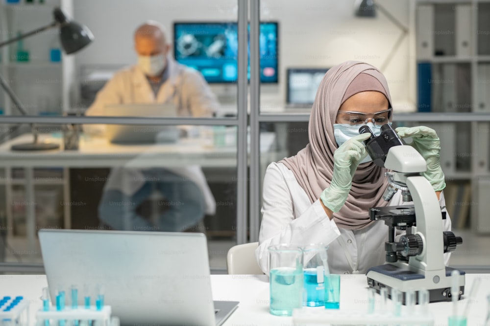 Young Muslim female virologist studying characteristics of new virus in microscope against male colleague networking in laboratory