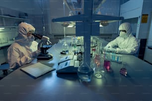 Due colleghi in abbigliamento da lavoro protettivo seduti al tavolo e che lavorano con campioni chimici in provette nel laboratorio
