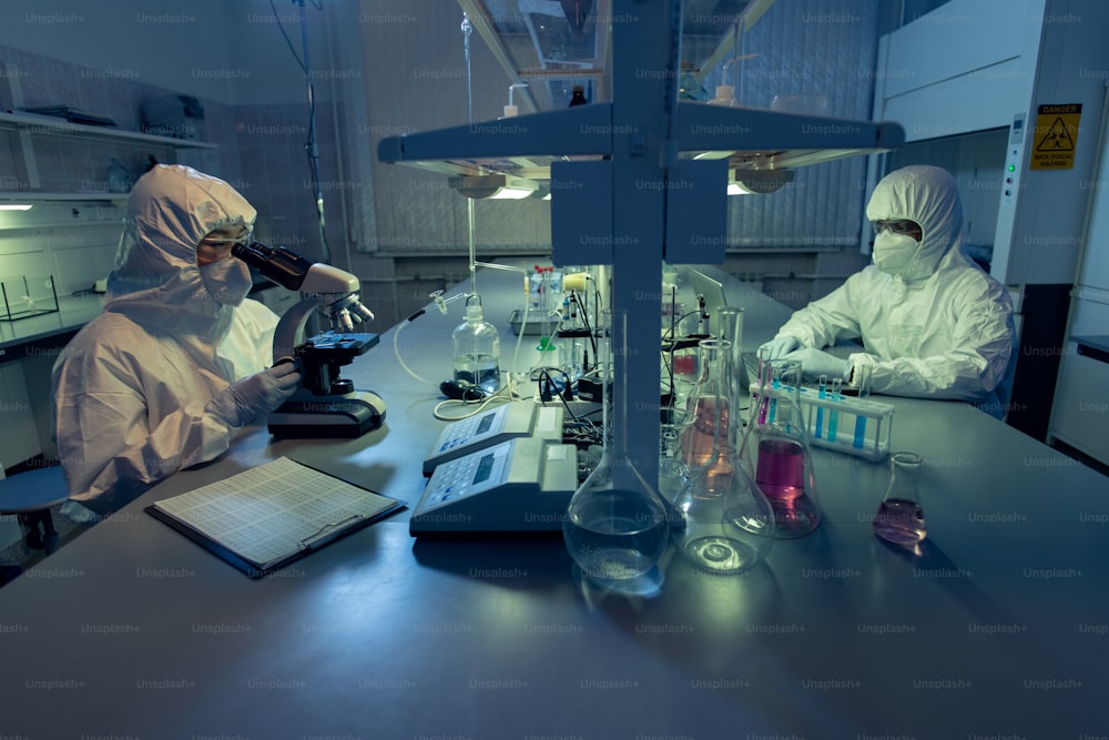 보호 작업복을 입은 두 명의 동료가 테이블에 앉아 실험실의 시험관에서 화학 샘플을 가지고 작업하고 있습니다.