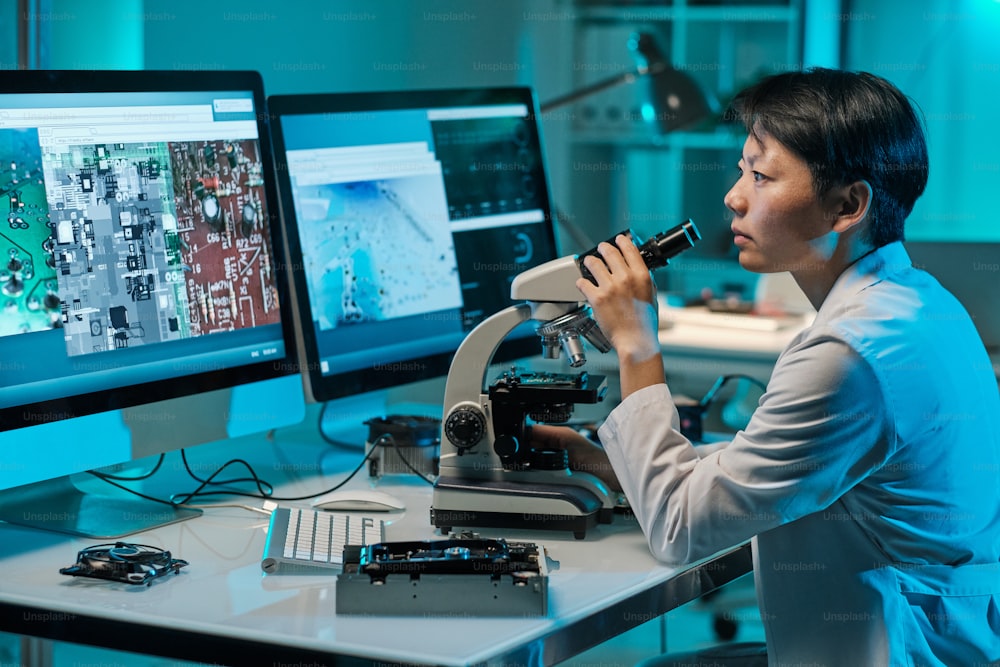 Young serious female scientist with microscope looking at computer screen while sitting by workplace