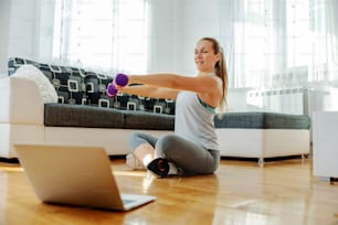 Strong sportswoman sitting on the floor at home and lifting dumbbells. She is following online tutorials.